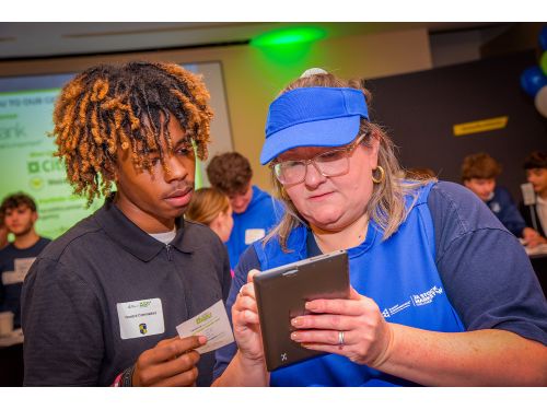 Students and volunteer engaging at the JA Stock Market Challenge; screen in background with sponsor information