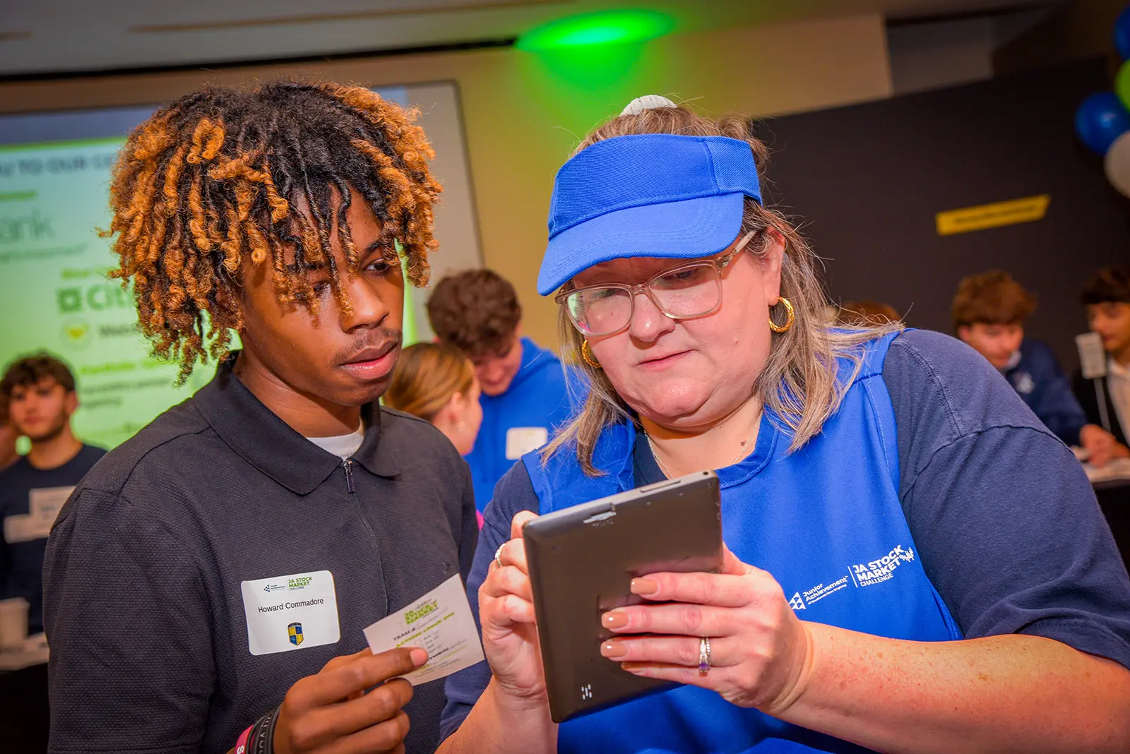 Students and volunteer engaging at the JA Stock Market Challenge; screen in background with sponsor information
