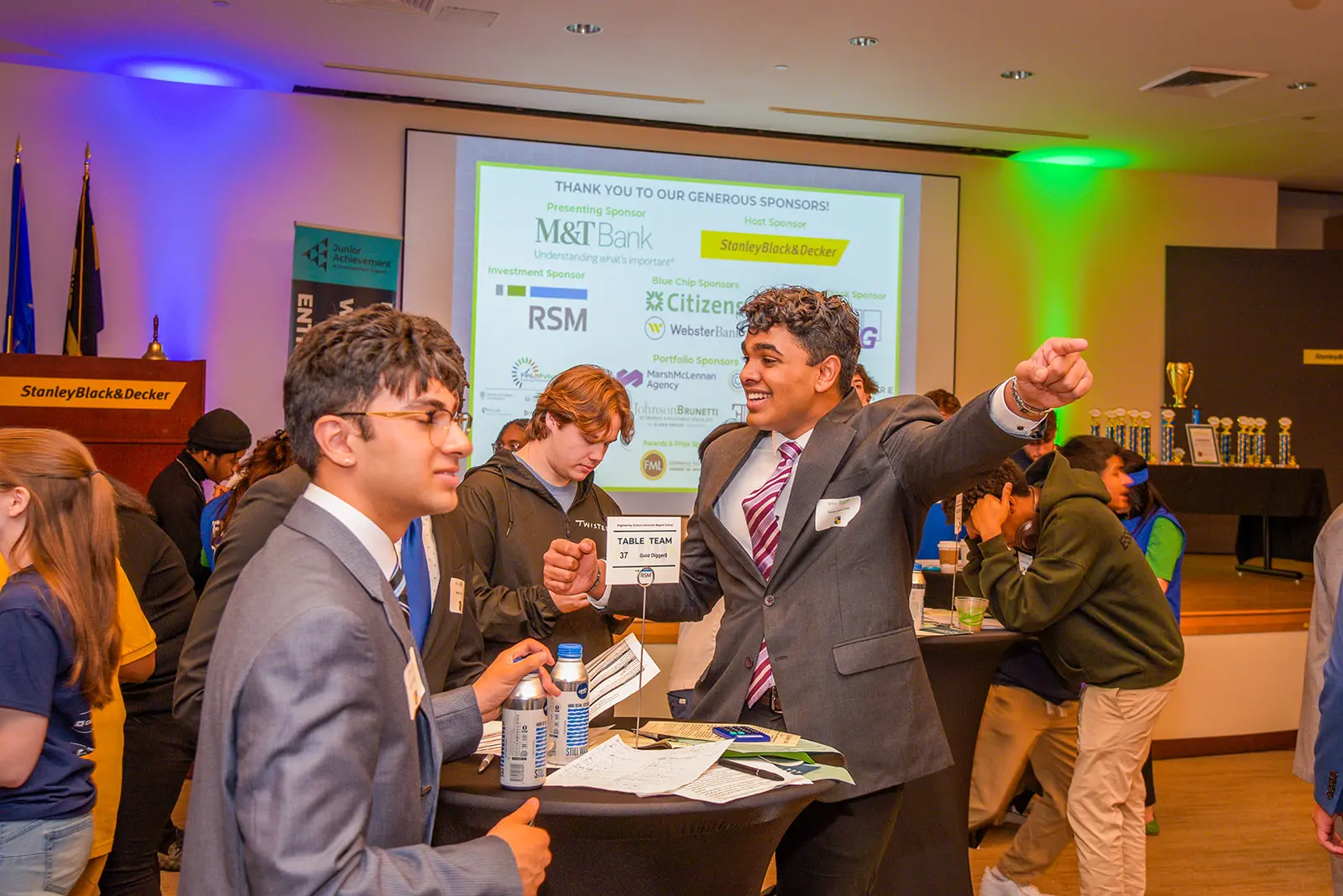 Students and volunteer engaging at the JA Stock Market Challenge; screen in background with sponsor information